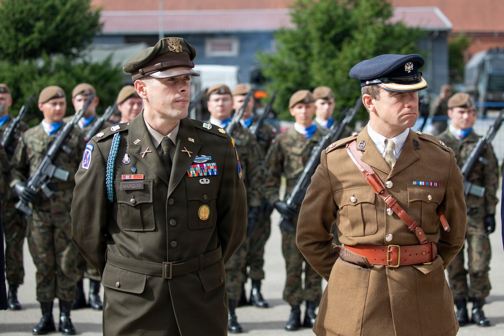 A military ceremony or parade featuring two senior officers in dress uniforms standing at attention in the foreground. The officer on the left wears a US Army dress uniform with the name tag 'MARQUIS' and multiple decorations, while the officer on the right wears what appears to be a British Army dress uniform with a Sam Browne belt. Behind them stands a formation of soldiers in combat uniforms carrying rifles.