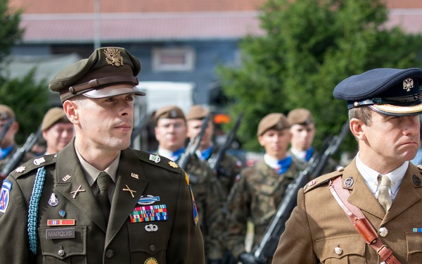 A military ceremony or parade featuring two senior officers in dress uniforms standing at attention in the foreground. The officer on the left wears a US Army dress uniform with the name tag 'MARQUIS' and multiple decorations, while the officer on the right wears what appears to be a British Army dress uniform with a Sam Browne belt. Behind them stands a formation of soldiers in combat uniforms carrying rifles.