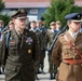 A military ceremony or parade featuring two senior officers in dress uniforms standing at attention in the foreground. The officer on the left wears a US Army dress uniform with the name tag 'MARQUIS' and multiple decorations, while the officer on the right wears what appears to be a British Army dress uniform with a Sam Browne belt. Behind them stands a formation of soldiers in combat uniforms carrying rifles.