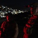 Two military personnel in full combat gear are positioned at an elevated outdoor location at night, illuminated by red light. A densely lit urban area is visible in the background below them. The foreground soldier wears a helmet-mounted night vision device and tactical vest.