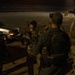 A nighttime scene showing military personnel and a law enforcement officer gathered in what appears to be an outdoor area near a barrier wall with concertina wire visible in the background. The group of approximately five individuals are engaged in a briefing or coordination discussion. The setting and equipment suggest a border security or tactical operation.