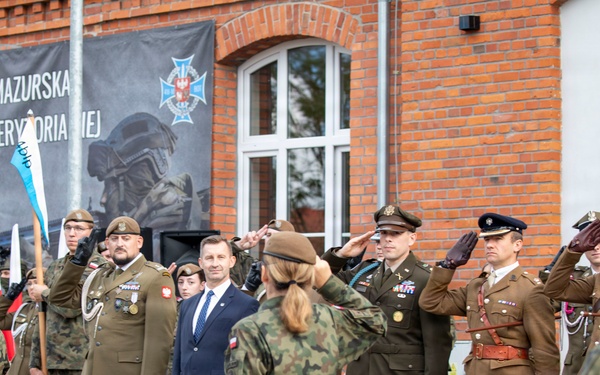 A military ceremony is taking place outside a brick building bearing a banner for '4 Warmińsko-Mazurska Brygada Obrony Terytorialnej' (4th Warmia-Mazury Territorial Defence Brigade) in Poland. Multiple military personnel in various uniforms, including Polish and what appear to be American officers, are rendering salutes during a formal review or parade. A civilian in a dark suit stands among the uniformed personnel, likely a government official.