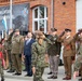 A military ceremony is taking place outside a brick building bearing a banner for '4 Warmińsko-Mazurska Brygada Obrony Terytorialnej' (4th Warmia-Mazury Territorial Defence Brigade) in Poland. Multiple military personnel in various uniforms, including Polish and what appear to be American officers, are rendering salutes during a formal review or parade. A civilian in a dark suit stands among the uniformed personnel, likely a government official.