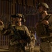 Two armed military personnel in full combat gear are positioned at night in front of a tall metal barrier fence topped with concertina/razor wire. The soldier on the left is equipped with night vision goggles mounted on his helmet and is carrying a rifle while pointing in a direction. Additional personnel are visible in the background near the fence.
