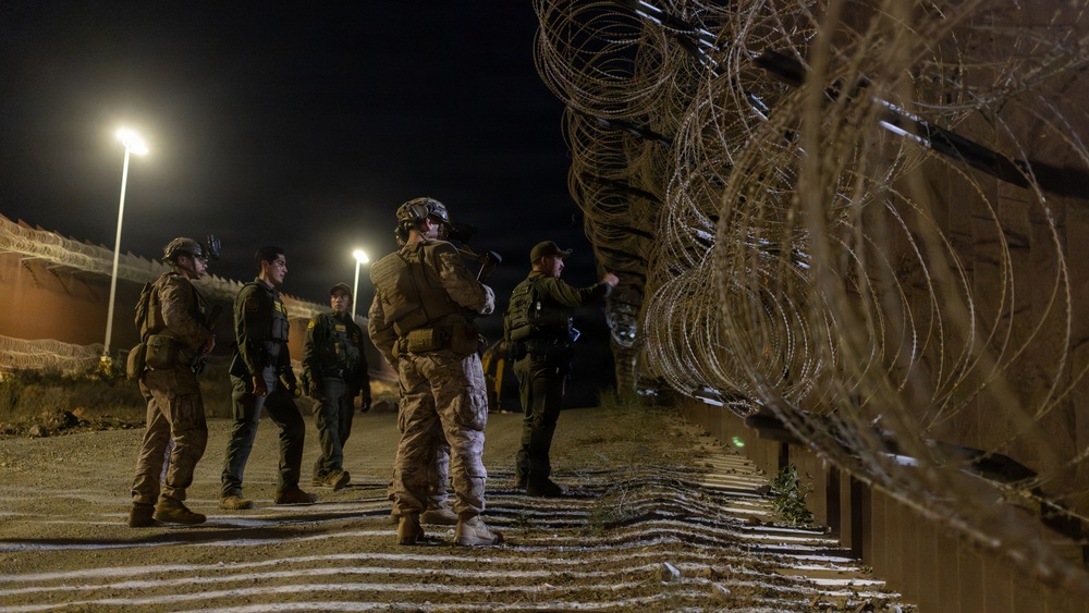 A nighttime scene showing uniformed personnel standing near a border barrier topped with concertina/razor wire. Multiple individuals in tactical gear and uniforms are gathered in a group alongside a metal fence structure. Artificial lighting from overhead poles illuminates the scene.