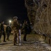 A nighttime scene showing uniformed personnel standing near a border barrier topped with concertina/razor wire. Multiple individuals in tactical gear and uniforms are gathered in a group alongside a metal fence structure. Artificial lighting from overhead poles illuminates the scene.