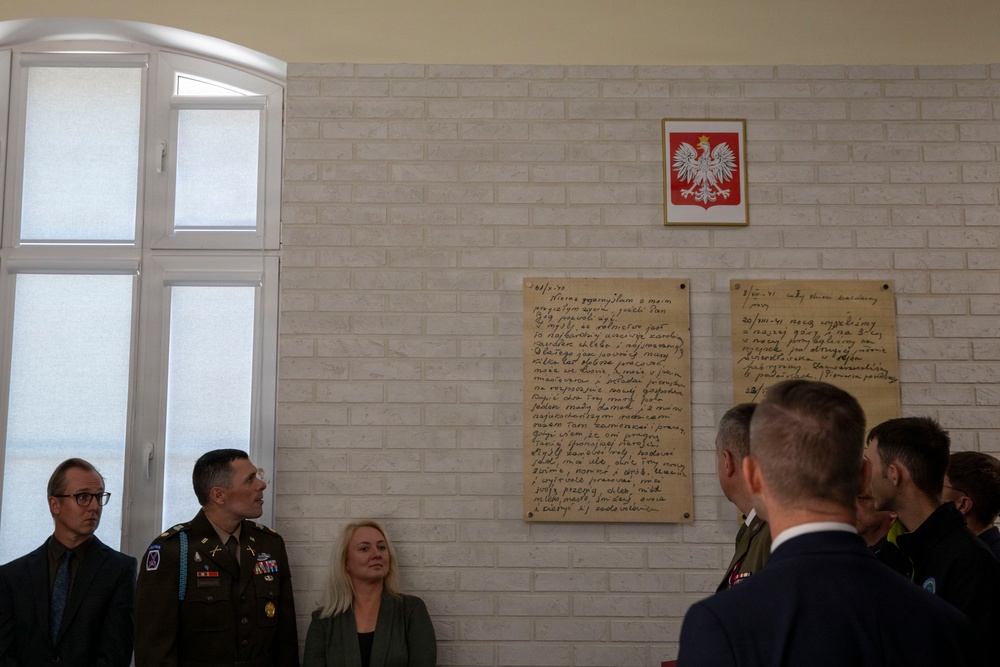 A group of military and civilian personnel are gathered in what appears to be a Polish institutional building, viewing commemorative plaques mounted on a white brick wall. Above the plaques hangs the coat of arms of Poland (white eagle on red background). The setting suggests an official ceremony or memorial event.