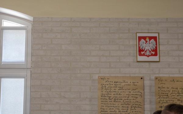 A group of military and civilian personnel are gathered in what appears to be a Polish institutional building, viewing commemorative plaques mounted on a white brick wall. Above the plaques hangs the coat of arms of Poland (white eagle on red background). The setting suggests an official ceremony or memorial event.