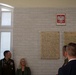 A group of military and civilian personnel are gathered in what appears to be a Polish institutional building, viewing commemorative plaques mounted on a white brick wall. Above the plaques hangs the coat of arms of Poland (white eagle on red background). The setting suggests an official ceremony or memorial event.