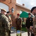 A group of multinational military personnel are gathered outside a brick building marked with the number '44', likely at a NATO or allied forces installation in Poland. Personnel from at least three nations are visible, including Polish, British, and American military officers in their respective uniforms. The gathering appears to be an informal meeting or reception event.