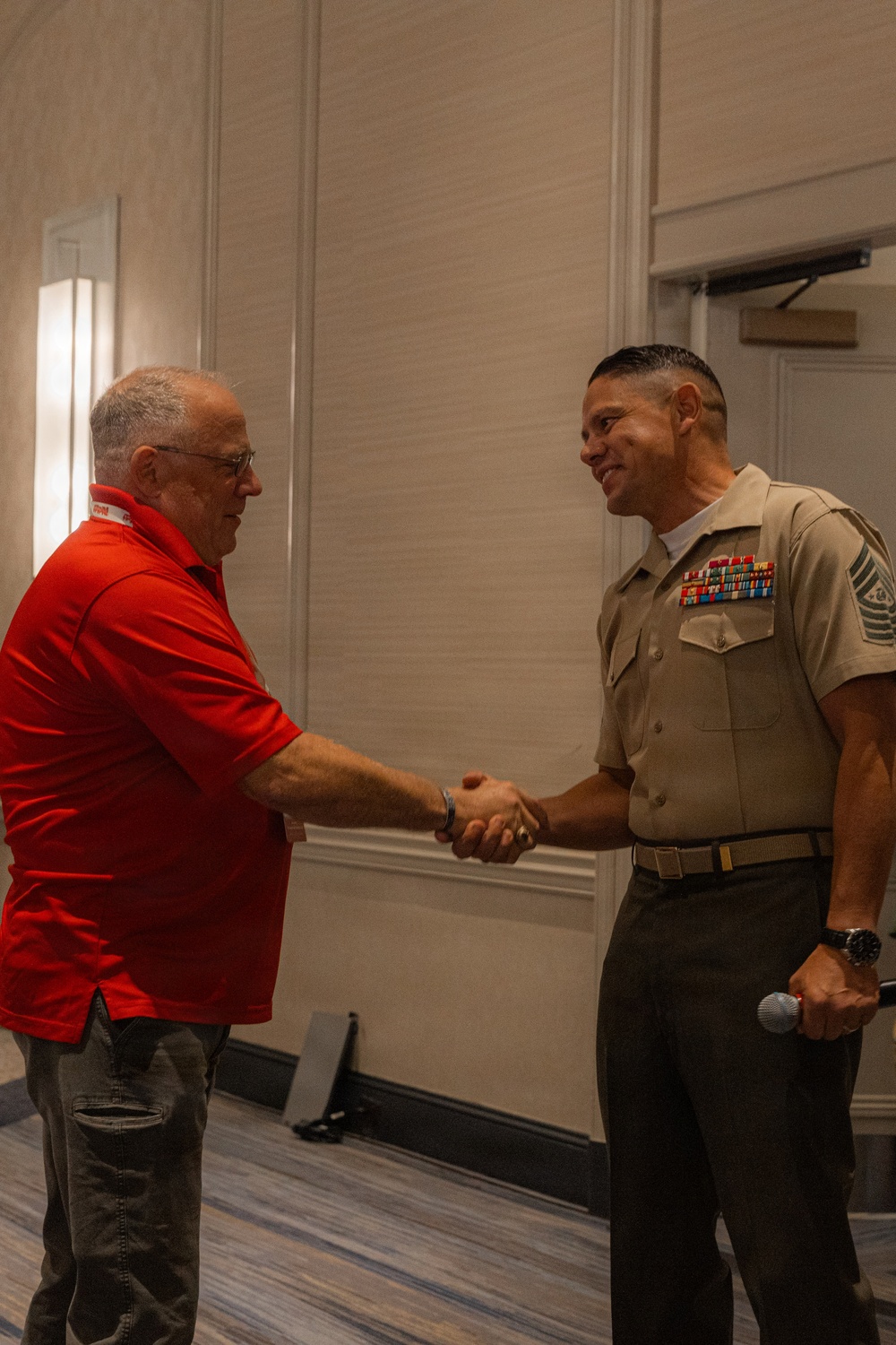 Two men are shaking hands in what appears to be a hotel ballroom or conference facility. One man is wearing a U.S. Marine Corps Service 'C' uniform with multiple service ribbons and senior enlisted rank insignia, while the other is dressed in a red polo shirt. The military figure is holding a microphone in his left hand, suggesting this may be a formal or ceremonial event.