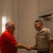 Two men are shaking hands in what appears to be a hotel ballroom or conference facility. One man is wearing a U.S. Marine Corps Service 'C' uniform with multiple service ribbons and senior enlisted rank insignia, while the other is dressed in a red polo shirt. The military figure is holding a microphone in his left hand, suggesting this may be a formal or ceremonial event.