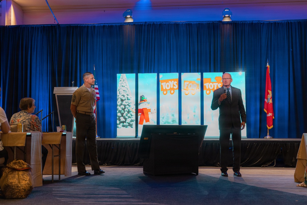 A formal event supporting the Toys for Tots charity program is taking place in a banquet or conference hall. Two men are on stage — one in U.S. Marine Corps dress uniform and one in a dark business suit holding a microphone — appearing to address an audience. The stage is decorated with a blue curtain backdrop, a Toys for Tots digital display, an American flag, and a Marine Corps flag.