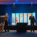 A formal event supporting the Toys for Tots charity program is taking place in a banquet or conference hall. Two men are on stage — one in U.S. Marine Corps dress uniform and one in a dark business suit holding a microphone — appearing to address an audience. The stage is decorated with a blue curtain backdrop, a Toys for Tots digital display, an American flag, and a Marine Corps flag.