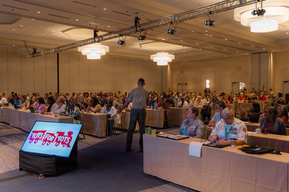 A large conference or meeting is taking place in a hotel ballroom with hundreds of attendees seated at long tables. A presenter stands at the front addressing the audience, with a 'Toys for Tots' branded display screen visible in the foreground. The event appears to be an official Toys for Tots organizational gathering, with some attendees in military or uniform attire.