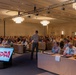 A large conference or meeting is taking place in a hotel ballroom with hundreds of attendees seated at long tables. A presenter stands at the front addressing the audience, with a 'Toys for Tots' branded display screen visible in the foreground. The event appears to be an official Toys for Tots organizational gathering, with some attendees in military or uniform attire.