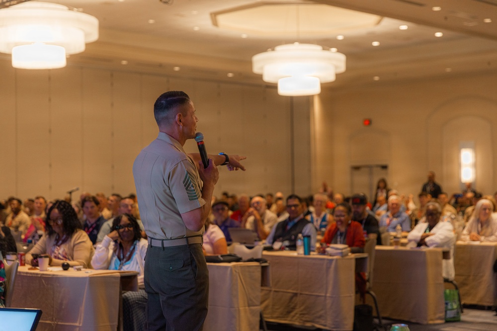 A military personnel in uniform is addressing a large audience in what appears to be a hotel ballroom or conference center. The speaker, holding a handheld microphone, is facing a crowd of approximately 50-100 attendees seated at round tables. The event appears to be a formal conference or symposium based on the venue setup and attendee lanyards.
