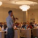 A military personnel in uniform is addressing a large audience in what appears to be a hotel ballroom or conference center. The speaker, holding a handheld microphone, is facing a crowd of approximately 50-100 attendees seated at round tables. The event appears to be a formal conference or symposium based on the venue setup and attendee lanyards.