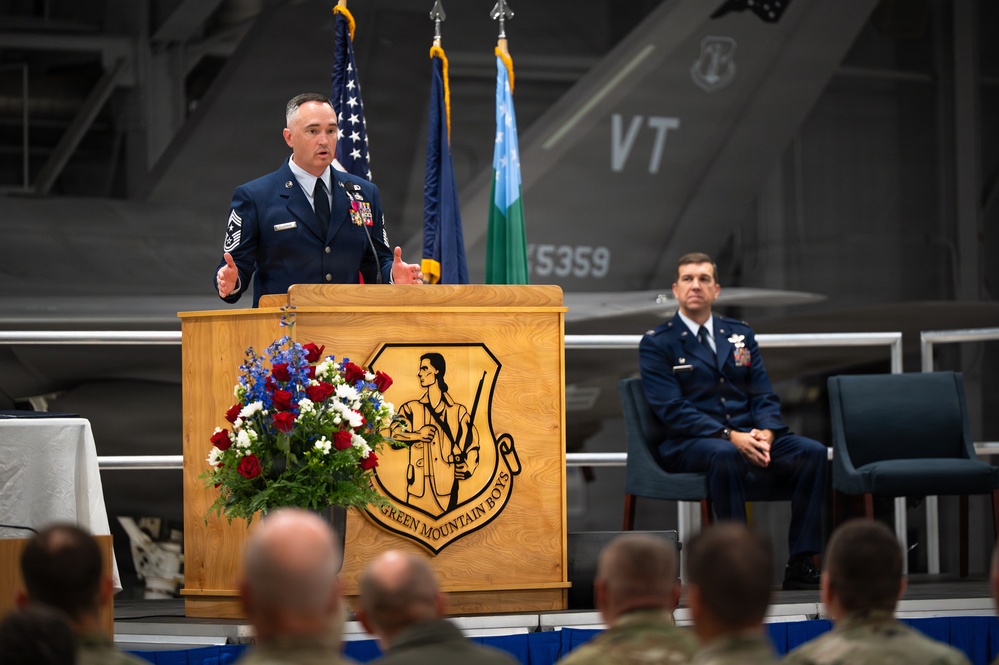 A senior enlisted military member in dress blue uniform delivers a speech at a podium bearing the 'Green Mountain Boys' insignia, likely at a formal military ceremony. The setting appears to be an aircraft hangar with a fighter jet visible in the background displaying 'VT' tail markings, consistent with the Vermont Air National Guard. A second uniformed officer is seated on stage behind the speaker, observing the proceedings.