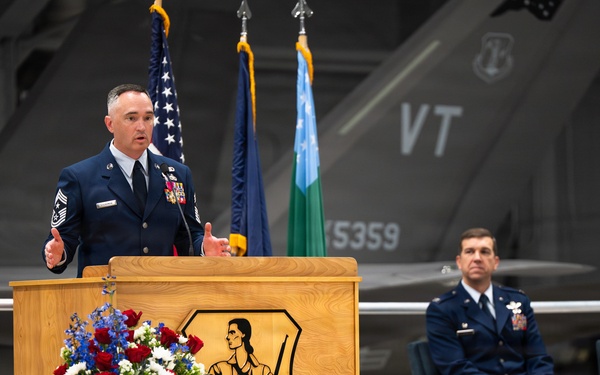 A senior enlisted military member in dress blue uniform delivers a speech at a podium bearing the 'Green Mountain Boys' insignia, likely at a formal military ceremony. The setting appears to be an aircraft hangar with a fighter jet visible in the background displaying 'VT' tail markings, consistent with the Vermont Air National Guard. A second uniformed officer is seated on stage behind the speaker, observing the proceedings.