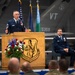 A senior enlisted military member in dress blue uniform delivers a speech at a podium bearing the 'Green Mountain Boys' insignia, likely at a formal military ceremony. The setting appears to be an aircraft hangar with a fighter jet visible in the background displaying 'VT' tail markings, consistent with the Vermont Air National Guard. A second uniformed officer is seated on stage behind the speaker, observing the proceedings.