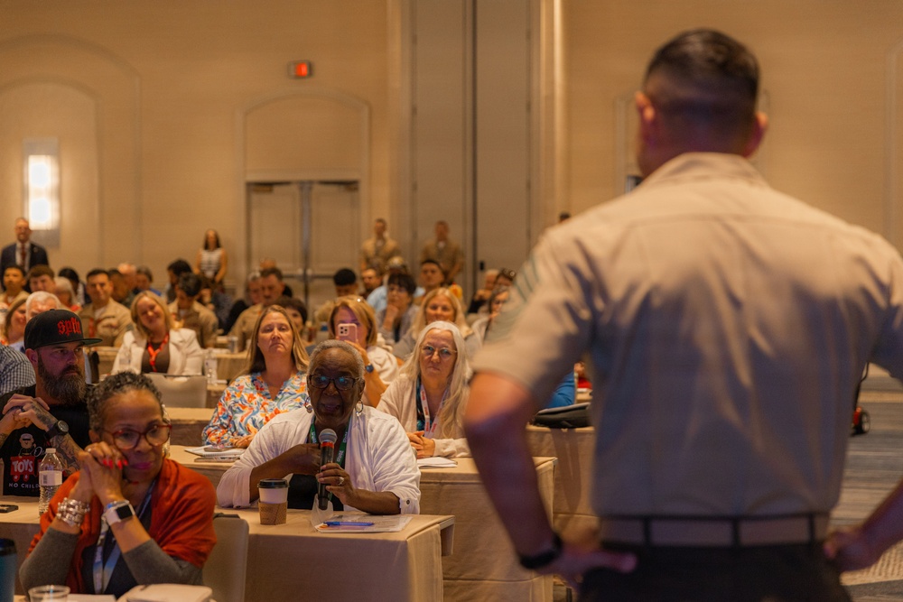 A conference or seminar setting in a large ballroom-style venue where an audience is seated at tables listening to a presenter. A woman in the foreground is holding a microphone, suggesting a Q&amp;A session or audience participation moment. The presenter, seen from behind, stands facing the crowd with hands on hips.