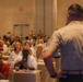 A conference or seminar setting in a large ballroom-style venue where an audience is seated at tables listening to a presenter. A woman in the foreground is holding a microphone, suggesting a Q&amp;A session or audience participation moment. The presenter, seen from behind, stands facing the crowd with hands on hips.