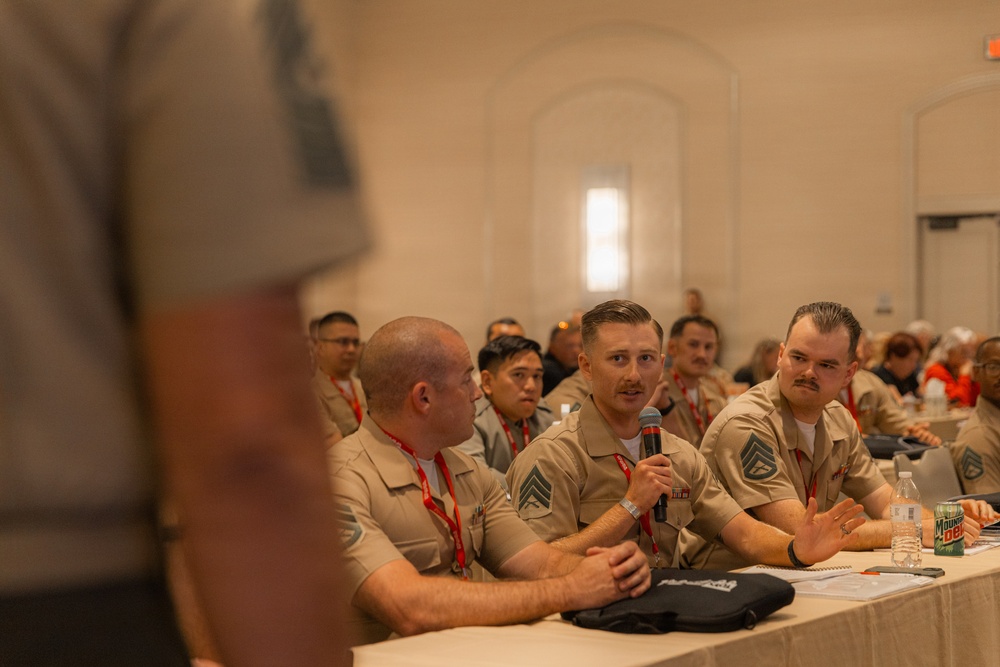A group of individuals wearing matching tan/khaki uniforms with rank insignia and red lanyards are seated at a table during what appears to be a formal meeting or conference. One individual in the center is speaking into a handheld microphone, suggesting a Q&amp;A session or panel discussion. The room is filled with similarly uniformed attendees seated in rows behind the main table.