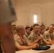 A group of individuals wearing matching tan/khaki uniforms with rank insignia and red lanyards are seated at a table during what appears to be a formal meeting or conference. One individual in the center is speaking into a handheld microphone, suggesting a Q&amp;A session or panel discussion. The room is filled with similarly uniformed attendees seated in rows behind the main table.