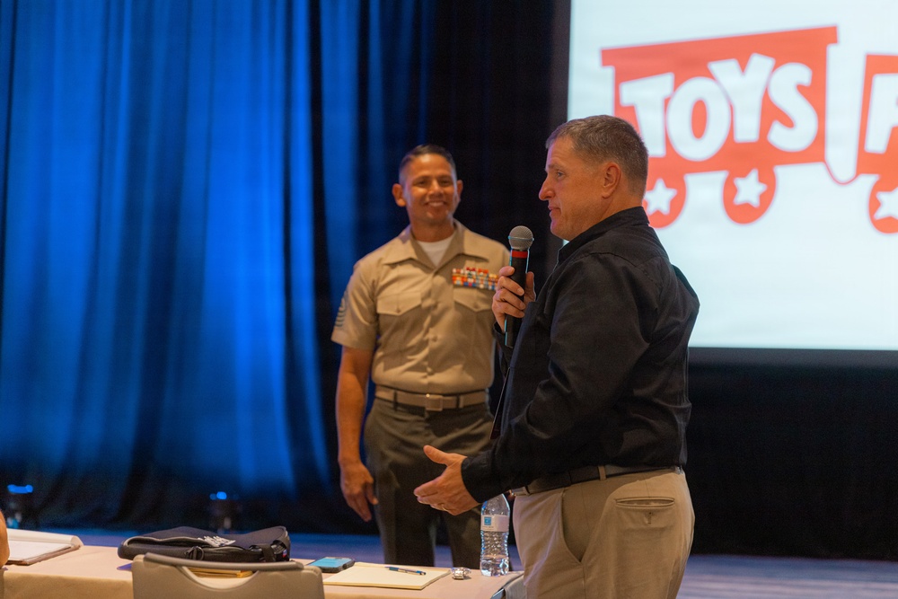 A man in a black jacket is speaking into a microphone at what appears to be a Toys for Tots event, with a U.S. Marine in dress uniform standing behind him smiling. The setting appears to be a conference or event hall with blue curtain draping and a projection screen displaying the Toys for Tots logo. The atmosphere is formal yet celebratory.