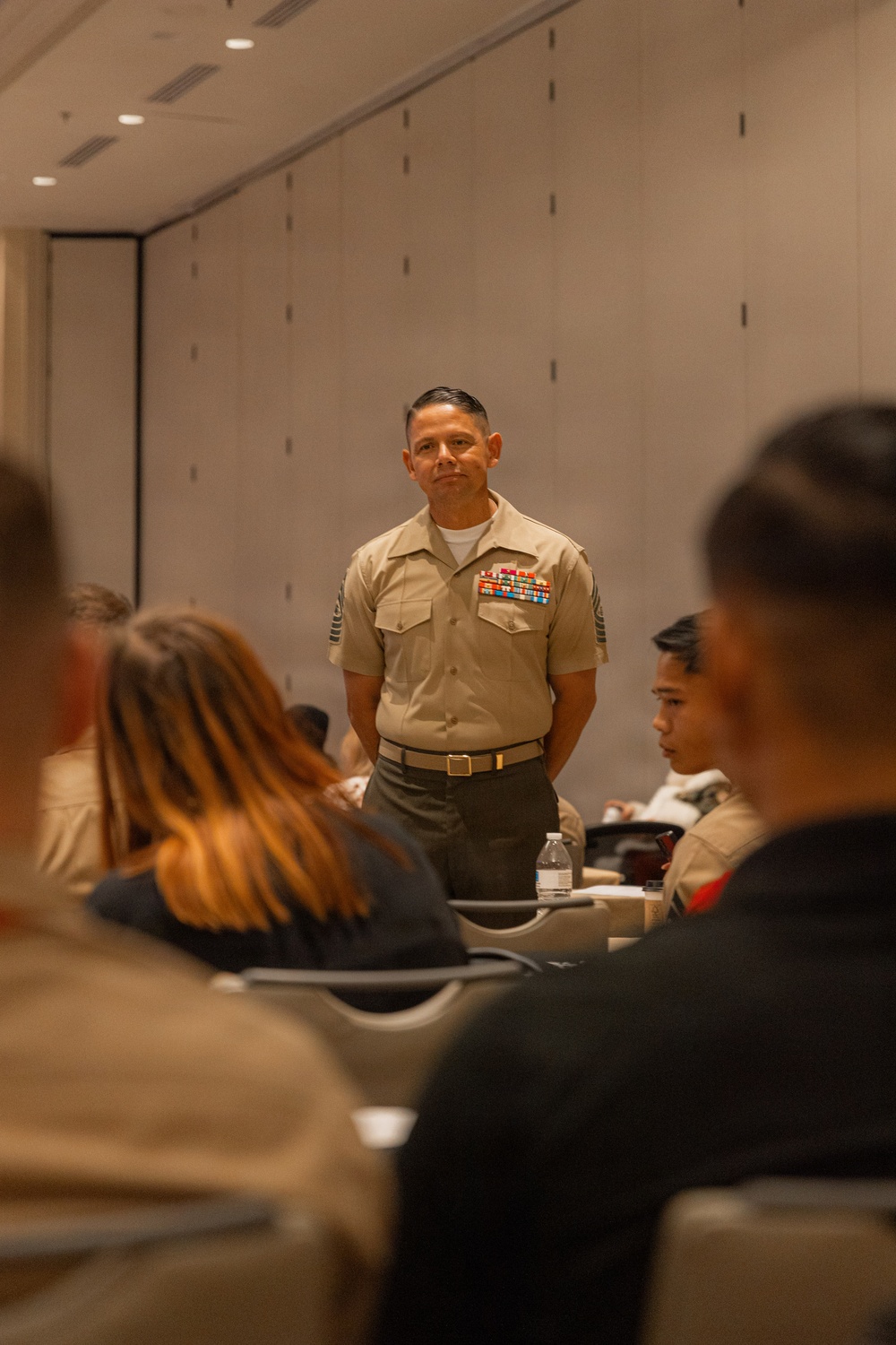 A senior U.S. Marine Corps non-commissioned officer in Service Charlie uniform stands at the front of a conference or briefing room, addressing a seated audience. He is wearing multiple rows of service ribbons and chevrons consistent with a senior enlisted rank (Sergeant Major or Master Sergeant). The setting appears to be a formal indoor briefing or training event.