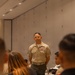A senior U.S. Marine Corps non-commissioned officer in Service Charlie uniform stands at the front of a conference or briefing room, addressing a seated audience. He is wearing multiple rows of service ribbons and chevrons consistent with a senior enlisted rank (Sergeant Major or Master Sergeant). The setting appears to be a formal indoor briefing or training event.