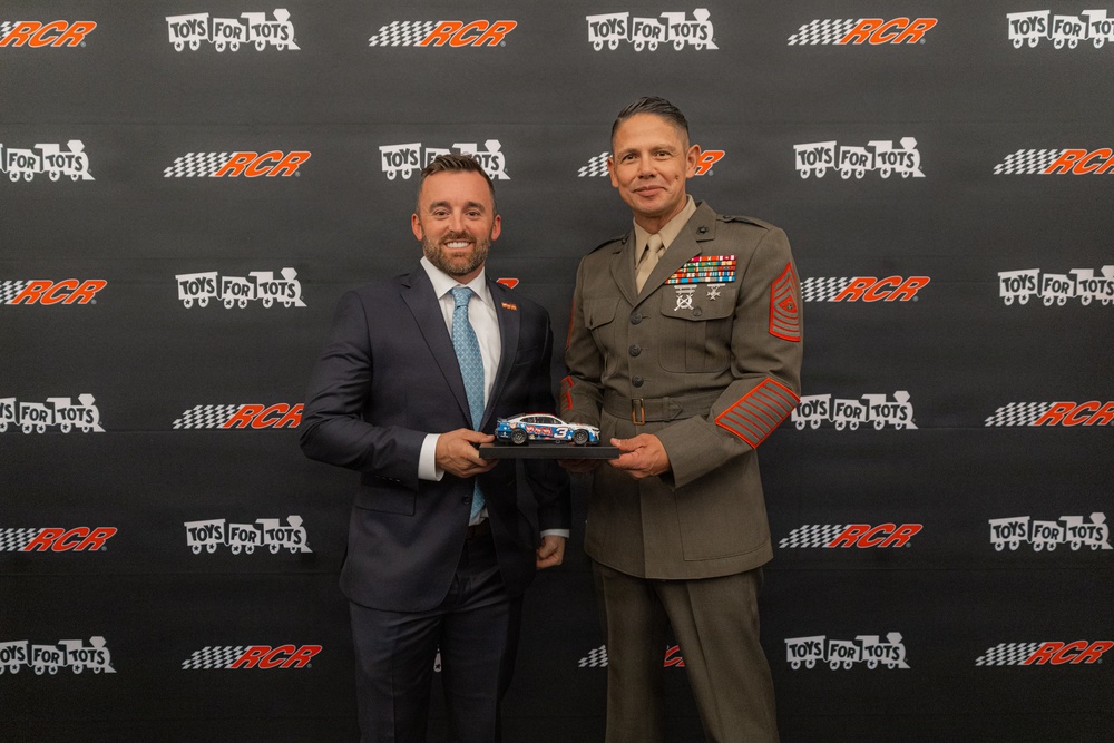 Two men pose together in front of a branded backdrop featuring RCR (Richard Childress Racing) and Toys for Tots logos. They are jointly holding a miniature die-cast NASCAR race car model. One man is in a business suit and the other is in a U.S. Marine Corps dress uniform with multiple service ribbons and sergeant major rank insignia.