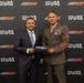Two men pose together in front of a branded backdrop featuring RCR (Richard Childress Racing) and Toys for Tots logos. They are jointly holding a miniature die-cast NASCAR race car model. One man is in a business suit and the other is in a U.S. Marine Corps dress uniform with multiple service ribbons and sergeant major rank insignia.