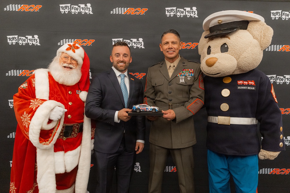 A promotional event photo featuring a partnership between RCR (Richard Childress Racing) and Toys for Tots. Two men — one in a business suit and one in a U.S. Marine Corps dress uniform — are holding a miniature NASCAR die-cast car together. They are flanked by a Santa Claus character on the left and the Toys for Tots mascot 'Gunny Bear' on the right.