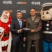 A promotional event photo featuring a partnership between RCR (Richard Childress Racing) and Toys for Tots. Two men — one in a business suit and one in a U.S. Marine Corps dress uniform — are holding a miniature NASCAR die-cast car together. They are flanked by a Santa Claus character on the left and the Toys for Tots mascot 'Gunny Bear' on the right.
