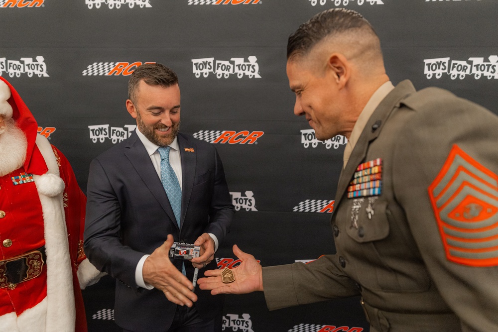A formal exchange is taking place at a Toys for Tots and RCR (Richard Childress Racing) event between a man in a navy suit and a U.S. Marine Corps Sergeant Major in dress uniform. The civilian appears to be presenting a small item, possibly a die-cast toy car, while the Marine holds what appears to be a challenge coin or rank insignia. A person dressed as Santa Claus is partially visible on the left side of the frame.