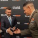 A formal exchange is taking place at a Toys for Tots and RCR (Richard Childress Racing) event between a man in a navy suit and a U.S. Marine Corps Sergeant Major in dress uniform. The civilian appears to be presenting a small item, possibly a die-cast toy car, while the Marine holds what appears to be a challenge coin or rank insignia. A person dressed as Santa Claus is partially visible on the left side of the frame.