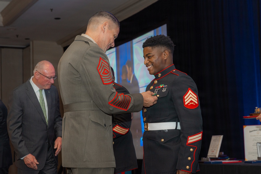 A formal U.S. Marine Corps ceremony is taking place in what appears to be a banquet or conference hall. A senior enlisted Marine in Service 'C' uniform (olive green) is pinning a medal or award onto a younger Marine wearing the dress blue uniform with Sergeant chevrons. The recipient is smiling broadly, suggesting this is a recognition or award ceremony.