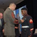 A formal U.S. Marine Corps ceremony is taking place in what appears to be a banquet or conference hall. A senior enlisted Marine in Service 'C' uniform (olive green) is pinning a medal or award onto a younger Marine wearing the dress blue uniform with Sergeant chevrons. The recipient is smiling broadly, suggesting this is a recognition or award ceremony.