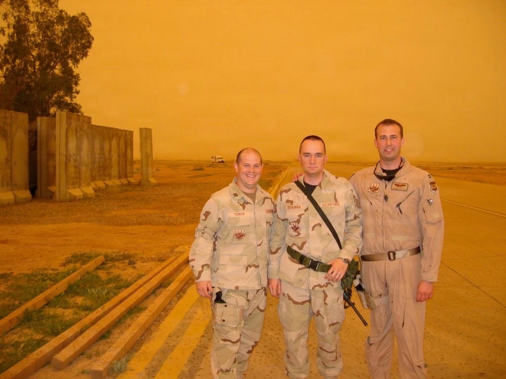Three U.S. military personnel pose for a photo outdoors during what appears to be a severe sandstorm, indicated by the heavy orange-tinted sky. Two individuals wear desert camouflage (DCU) uniforms while the third wears a tan flight suit. Name tapes visible on uniforms read 'TOMASI', 'STEADMAN', and 'U.S. AIR FORCE', suggesting an Air Force deployment likely in the Middle East.