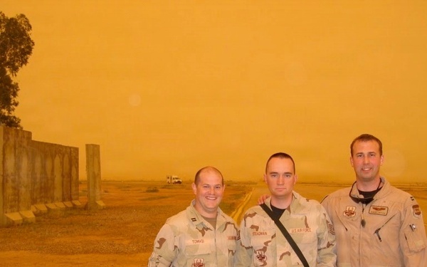 Three U.S. military personnel pose for a photo outdoors during what appears to be a severe sandstorm, indicated by the heavy orange-tinted sky. Two individuals wear desert camouflage (DCU) uniforms while the third wears a tan flight suit. Name tapes visible on uniforms read 'TOMASI', 'STEADMAN', and 'U.S. AIR FORCE', suggesting an Air Force deployment likely in the Middle East.