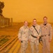 Three U.S. military personnel pose for a photo outdoors during what appears to be a severe sandstorm, indicated by the heavy orange-tinted sky. Two individuals wear desert camouflage (DCU) uniforms while the third wears a tan flight suit. Name tapes visible on uniforms read 'TOMASI', 'STEADMAN', and 'U.S. AIR FORCE', suggesting an Air Force deployment likely in the Middle East.