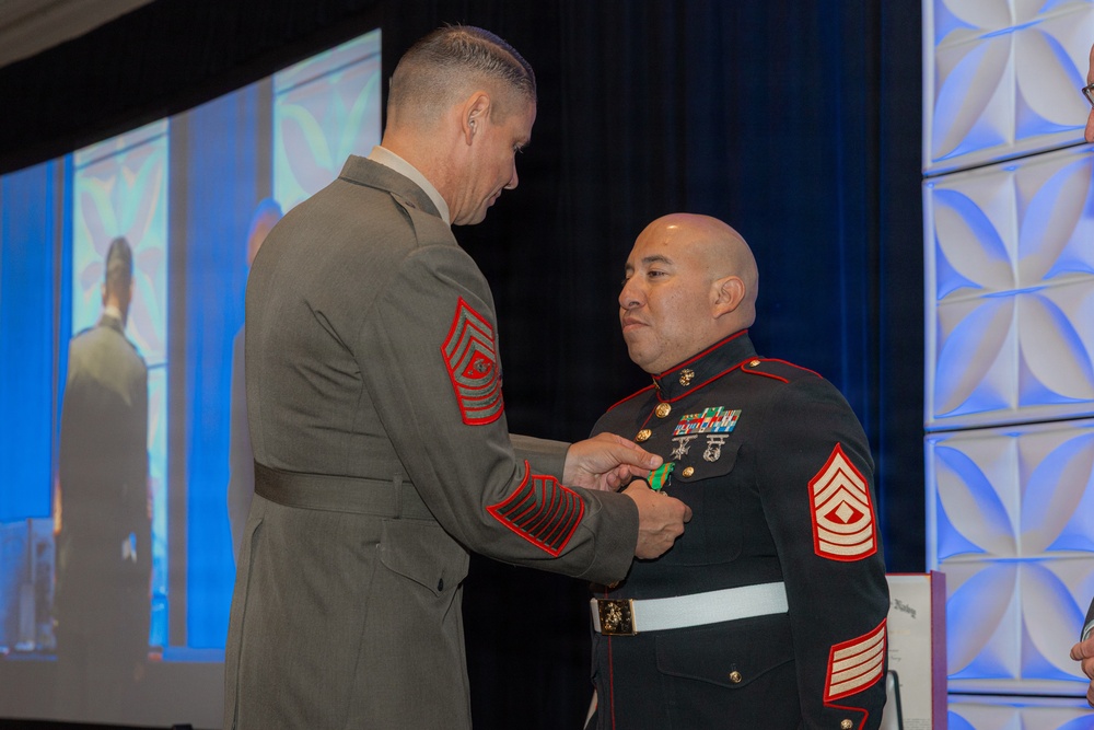 Two U.S. Marine Corps senior non-commissioned officers are participating in a formal awards or decoration ceremony at what appears to be an indoor event venue. The Marine on the left, wearing Service 'C' uniform with Sergeant Major chevrons, is pinning a medal or decoration onto the Marine on the right, who is wearing the Blue Dress uniform also with Master Gunnery Sergeant or Sergeant Major chevrons. A large projection screen is visible in the background displaying what appears to be the same ceremony.