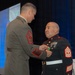 Two U.S. Marine Corps senior non-commissioned officers are participating in a formal awards or decoration ceremony at what appears to be an indoor event venue. The Marine on the left, wearing Service 'C' uniform with Sergeant Major chevrons, is pinning a medal or decoration onto the Marine on the right, who is wearing the Blue Dress uniform also with Master Gunnery Sergeant or Sergeant Major chevrons. A large projection screen is visible in the background displaying what appears to be the same ceremony.
