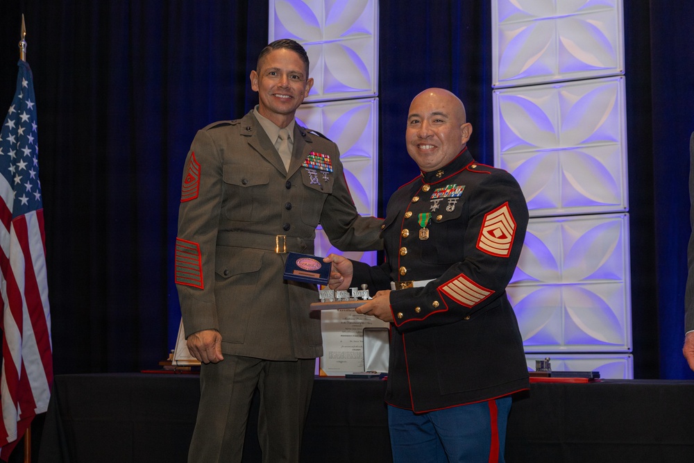 Two U.S. Marine Corps senior non-commissioned officers are shaking hands during what appears to be a formal awards or recognition ceremony. The Marine on the left is wearing the olive green 'Charlie' service uniform, while the Marine on the right is wearing the dress blue uniform. Both display the rank insignia of Master Gunnery Sergeant or Sergeant Major, and both have extensive medal and ribbon displays.