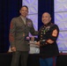 Two U.S. Marine Corps senior non-commissioned officers are shaking hands during what appears to be a formal awards or recognition ceremony. The Marine on the left is wearing the olive green 'Charlie' service uniform, while the Marine on the right is wearing the dress blue uniform. Both display the rank insignia of Master Gunnery Sergeant or Sergeant Major, and both have extensive medal and ribbon displays.