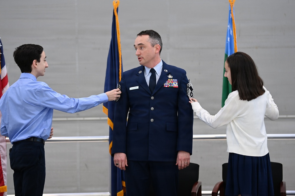 A U.S. Air Force service member in dress blues is having insignia or rank devices pinned onto his shoulders by two young people, likely family members, during a formal military promotion ceremony. The setting appears to be an official indoor venue with Air Force flags visible in the background. This type of ceremony traditionally involves family members placing new rank insignia on the service member.