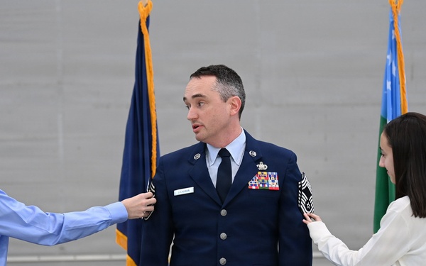 A U.S. Air Force service member in dress blues is having insignia or rank devices pinned onto his shoulders by two young people, likely family members, during a formal military promotion ceremony. The setting appears to be an official indoor venue with Air Force flags visible in the background. This type of ceremony traditionally involves family members placing new rank insignia on the service member.
