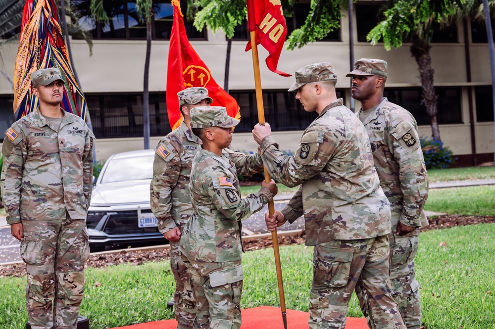 A U.S. Army change of command or activation ceremony is taking place outdoors, likely on a military installation in a tropical location. Two soldiers are exchanging a red unit guidon (flag marked 'HQ'), which is the central act of a formal military ceremony signifying transfer of command authority. Four soldiers in OCP (Operational Camouflage Pattern) uniforms are visible, with unit colors and a streamer-adorned organizational flag in the background.